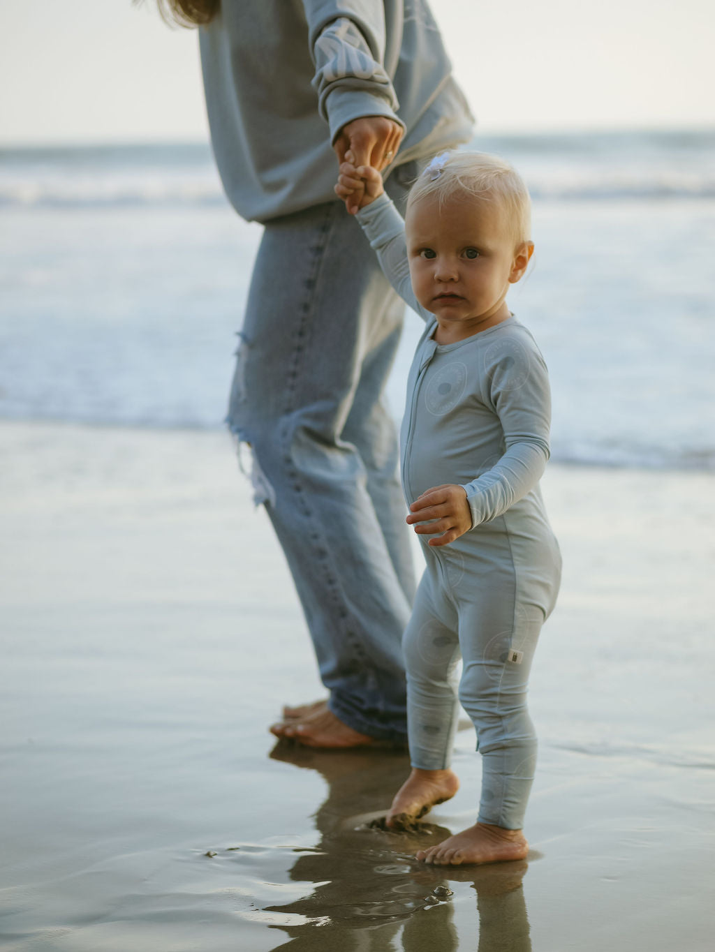 A toddler in forever french baby's hypo-allergenic Bamboo Zip Pajamas | San Clemente holds an adult's hand, standing barefoot on wet beach sand. The partially visible adult in ripped jeans stands with the ocean stretching out behind them.