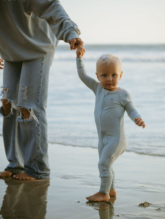 A toddler wearing forever french baby Bamboo Zip Pajamas | San Clemente holds an adult's hand while walking on wet beach sand. The adult is in ripped jeans and a sweatshirt, with the ocean in the background.