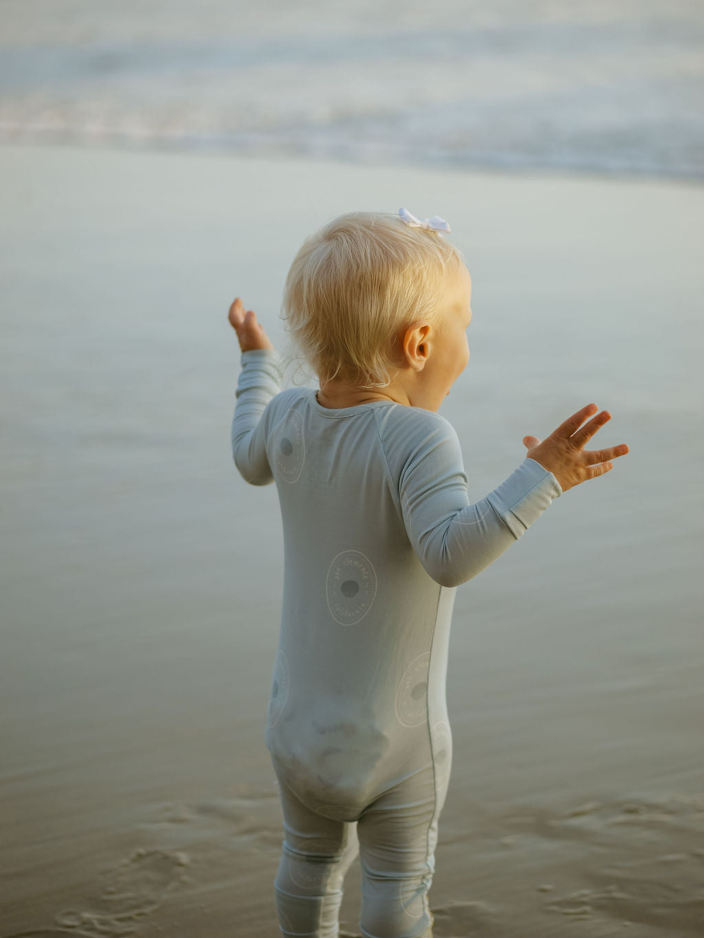 A young child wearing forever french baby Bamboo Zip Pajamas | San Clemente stands with arms raised at the ocean’s edge, facing the water and sandy shore.