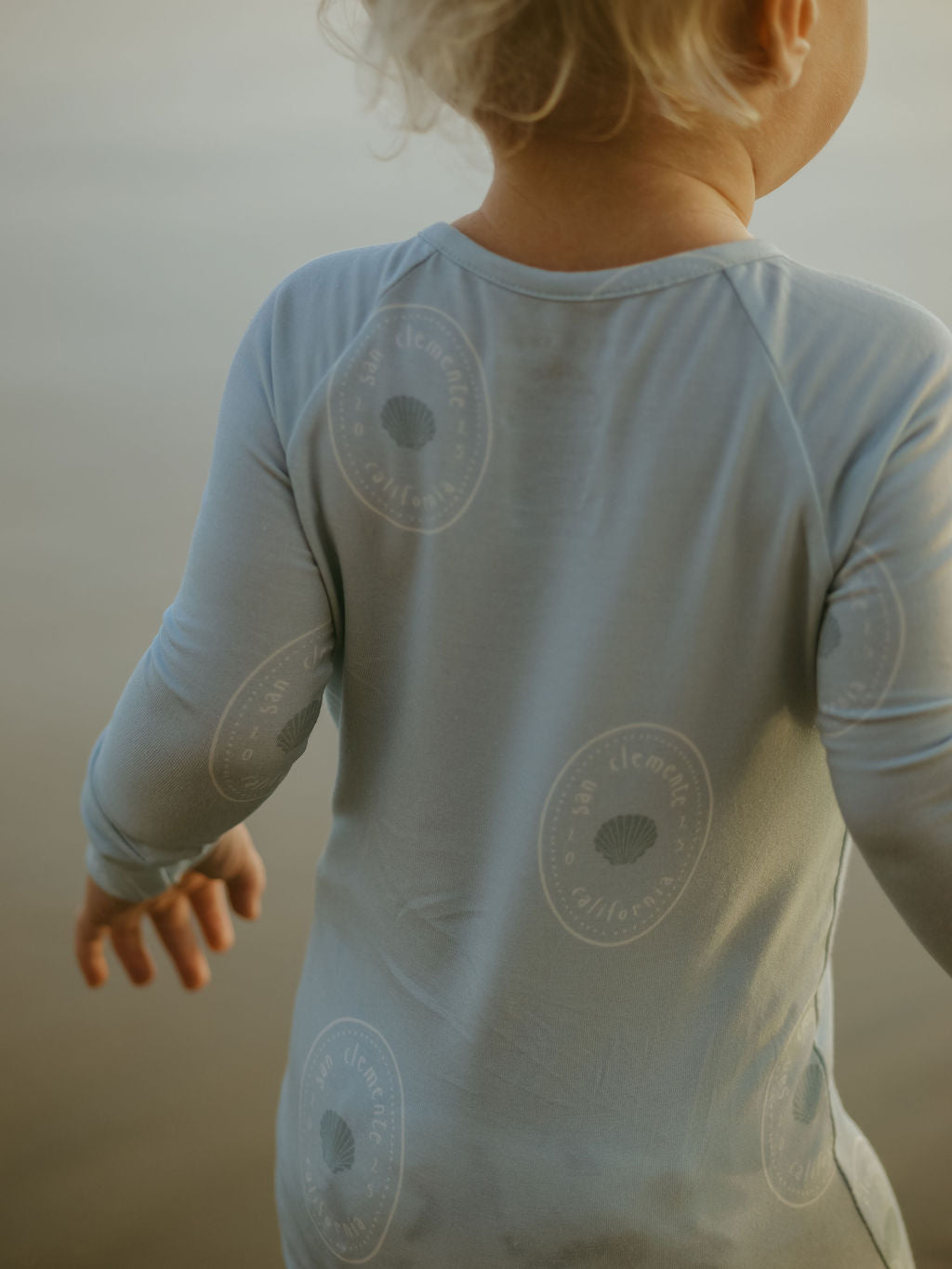 A young child with blonde hair, seen from behind, wears forever french baby Bamboo Zip Pajamas | San Clemente in light blue, featuring circular “San Clemente, California” logos and seashell graphics—hypo-allergenic for comfort.
