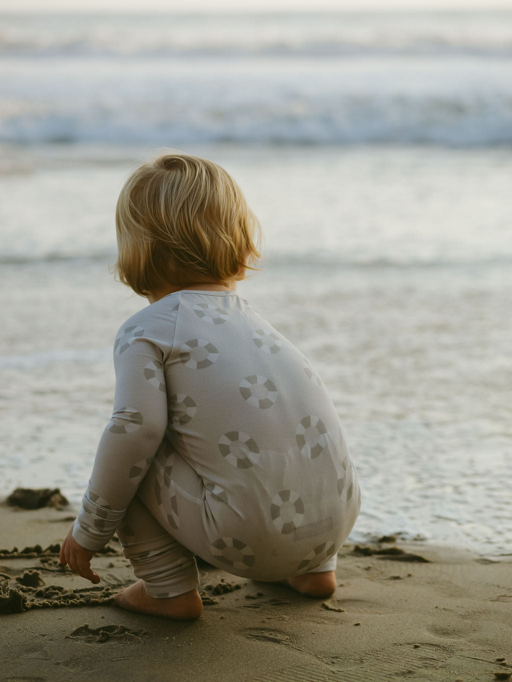 A young blonde child wears forever french baby's Bamboo Zip Pajamas | Lifesaver, squatting barefoot on the sandy beach and facing the ocean waves in soft light.