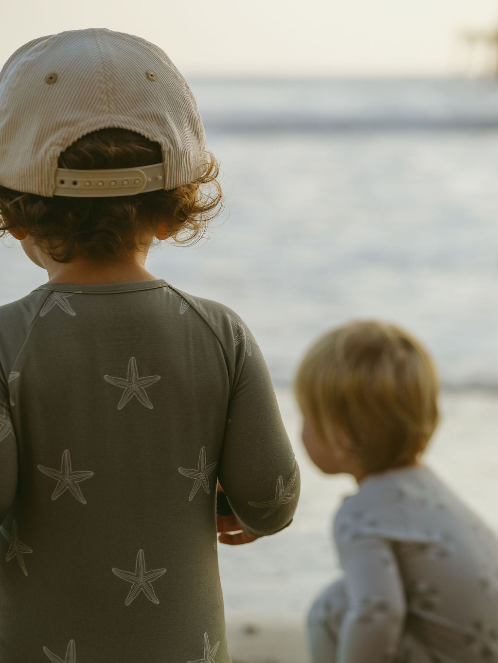 Two young children wear forever french baby Bamboo Zip Pajamas | Starfish as they play on the beach—one in a backward cap gazes at the waves, while the other crouches nearby, both highlighted by soft, warm light.