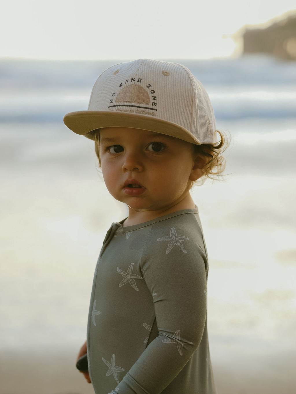 A young child wearing forever french baby's Bamboo Zip Pajamas | Starfish stands on a beach in soft, hypoallergenic starfish-patterned pajamas and cap, gazing at the camera with waves and a blurred shoreline behind them.