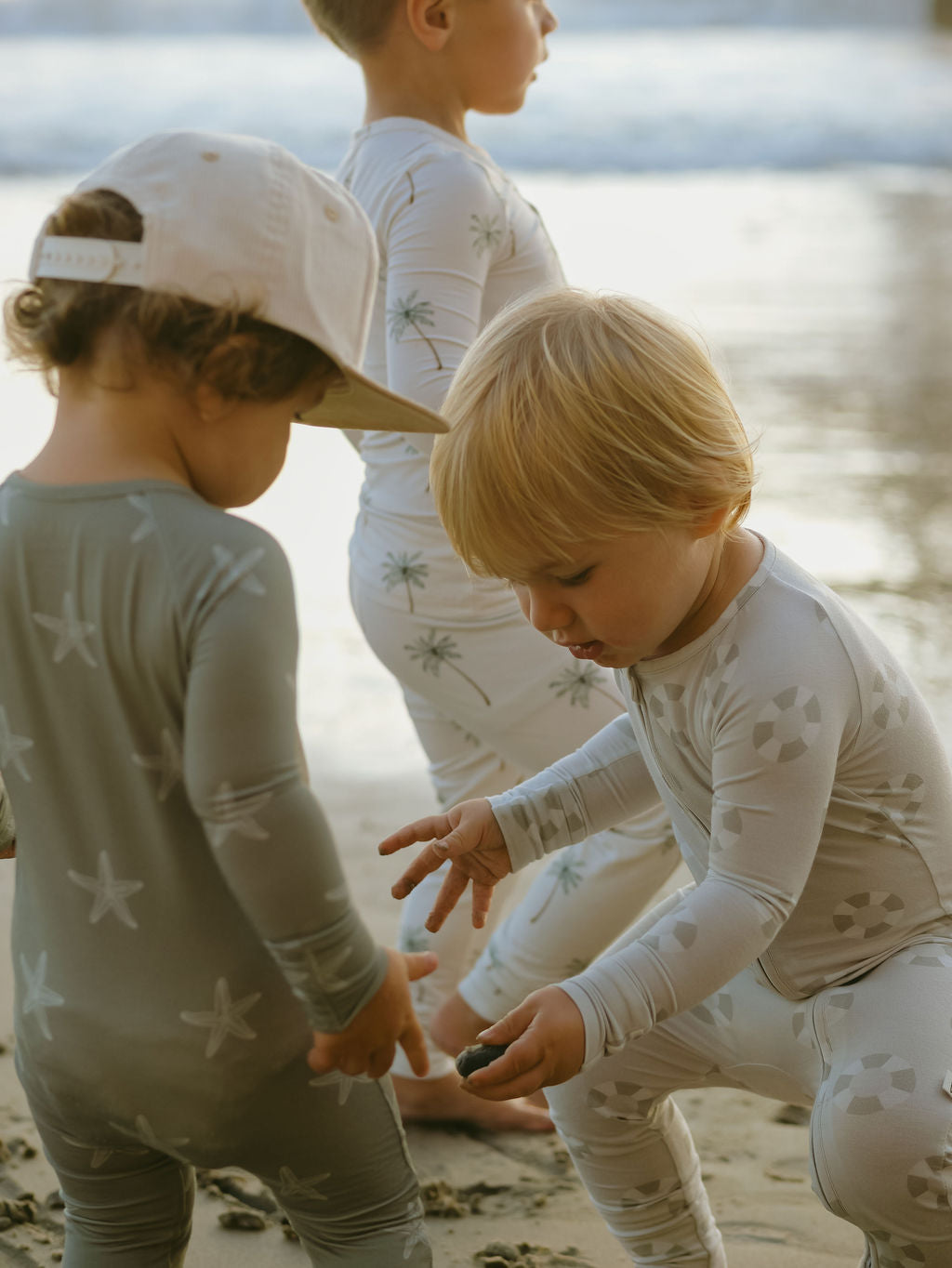 Three young children wear forever french baby’s Bamboo Zip Pajamas | Starfish, featuring playful patterns, as they stand and play on a sandy beach. One child in a cap looks down as another picks something up near the water.