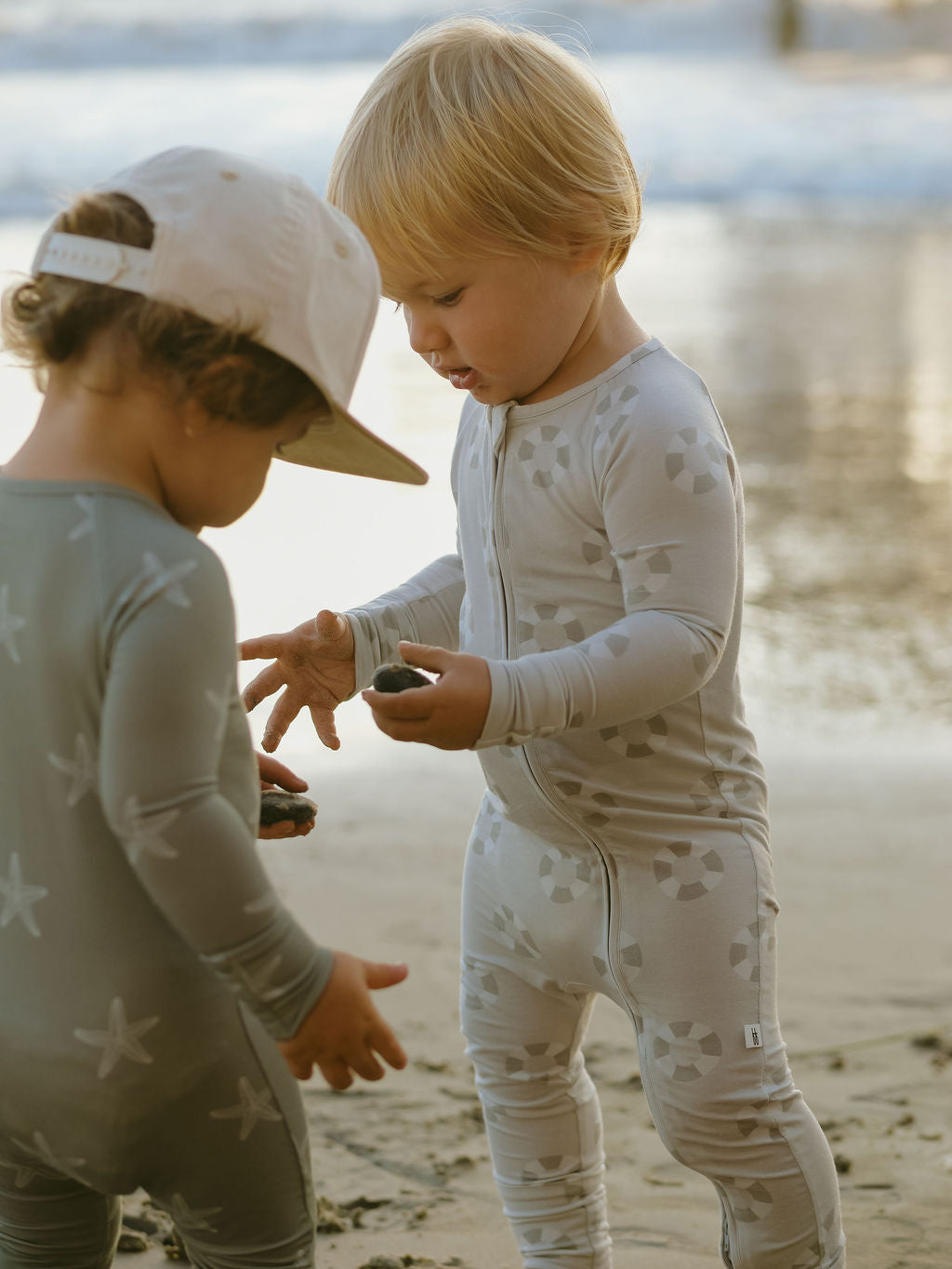 Two young children wear forever french baby Bamboo Zip Pajamas | Lifesaver on a sandy beach. One hands dark objects, possibly rocks or shells, to the other as the ocean and wet sand appear in the background.