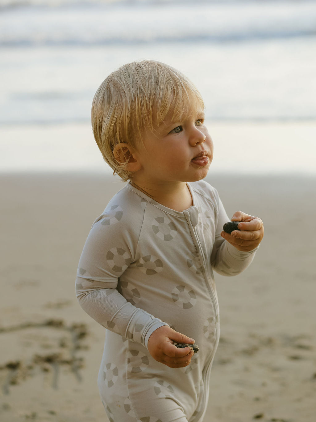 A young child with blonde hair wearing forever french baby Bamboo Zip Pajamas | Lifesaver, featuring a geometric pattern, stands on a sandy beach with small rocks in hand, as the ocean waves blur in the background.