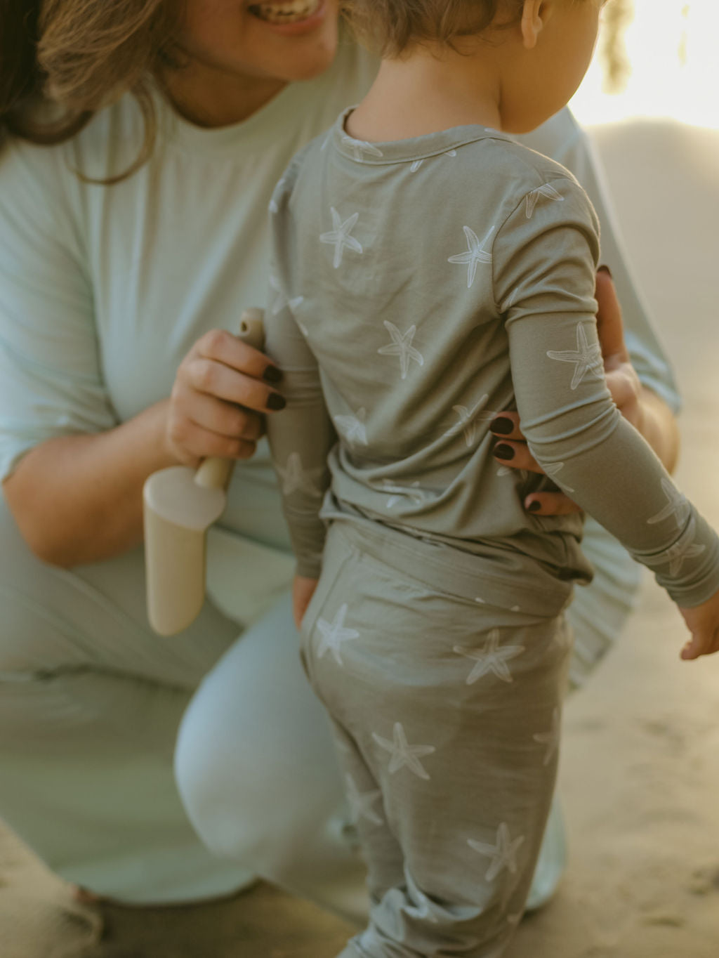 A woman smiles and kneels beside a child in forever french baby Bamboo Two Piece Pajamas | Starfish, holding a small beige toy shovel on sandy ground.