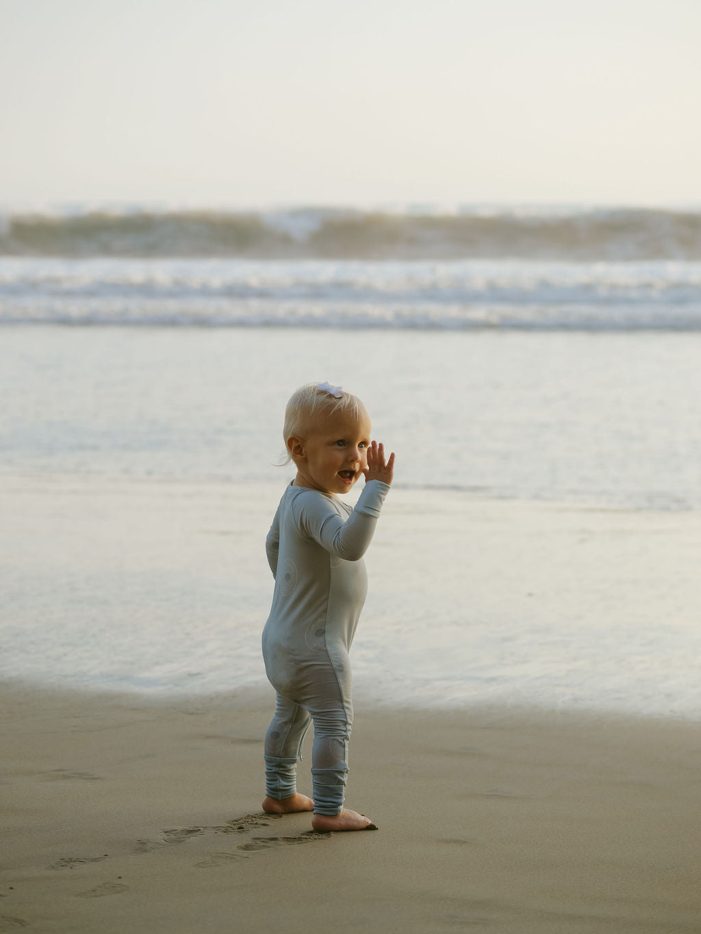 A young child in forever french baby's Bamboo Zip Pajamas | San Clemente stands barefoot at the beach’s edge, smiling and waving as gentle waves roll in behind.