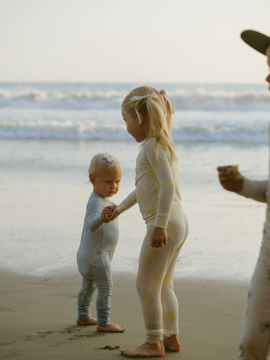 Two young children wearing forever french baby Bamboo Two Piece Pajamas in Lemon Drop stand barefoot by the water, holding hands as the older gently guides the younger. Waves and soft sunlight create a peaceful, dreamy beach scene.