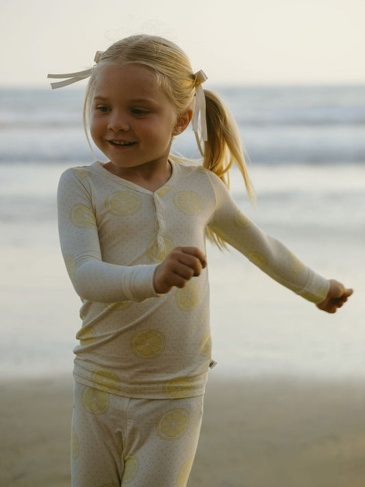 A smiling young girl with blond pigtails extends her arms on a beach at sunset, wearing forever french baby's Bamboo Two Piece Pajamas | Lemon Drop in white with yellow lemon prints.