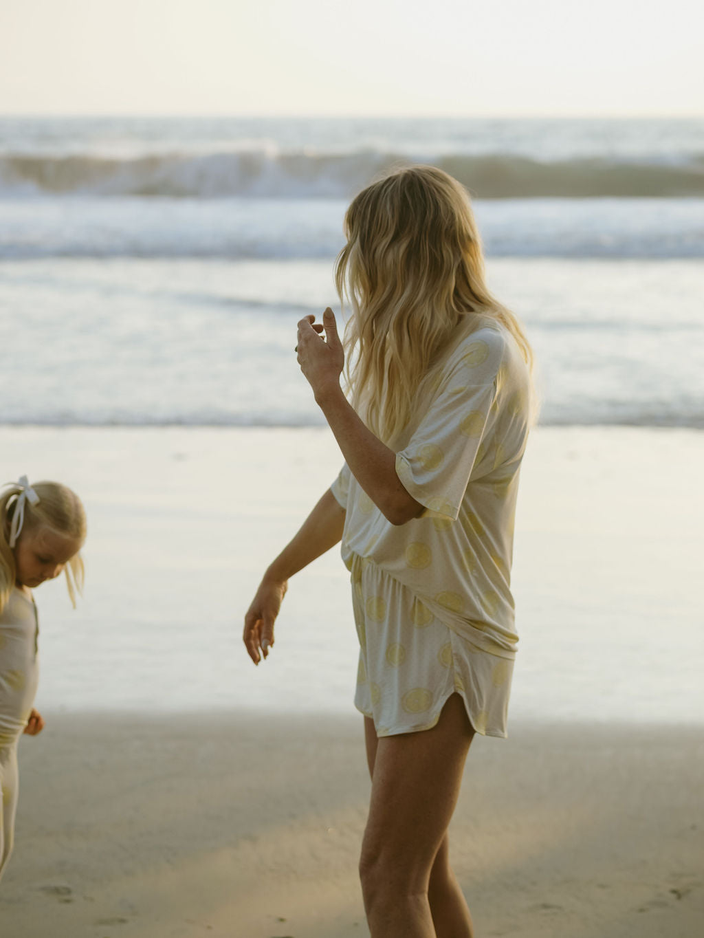 A woman with long blonde hair wearing the forever french baby Women's Bamboo Short Set in Lemon Drop stands on a beach at sunset, while a young girl with a hair ribbon walks nearby. Waves roll in the background.