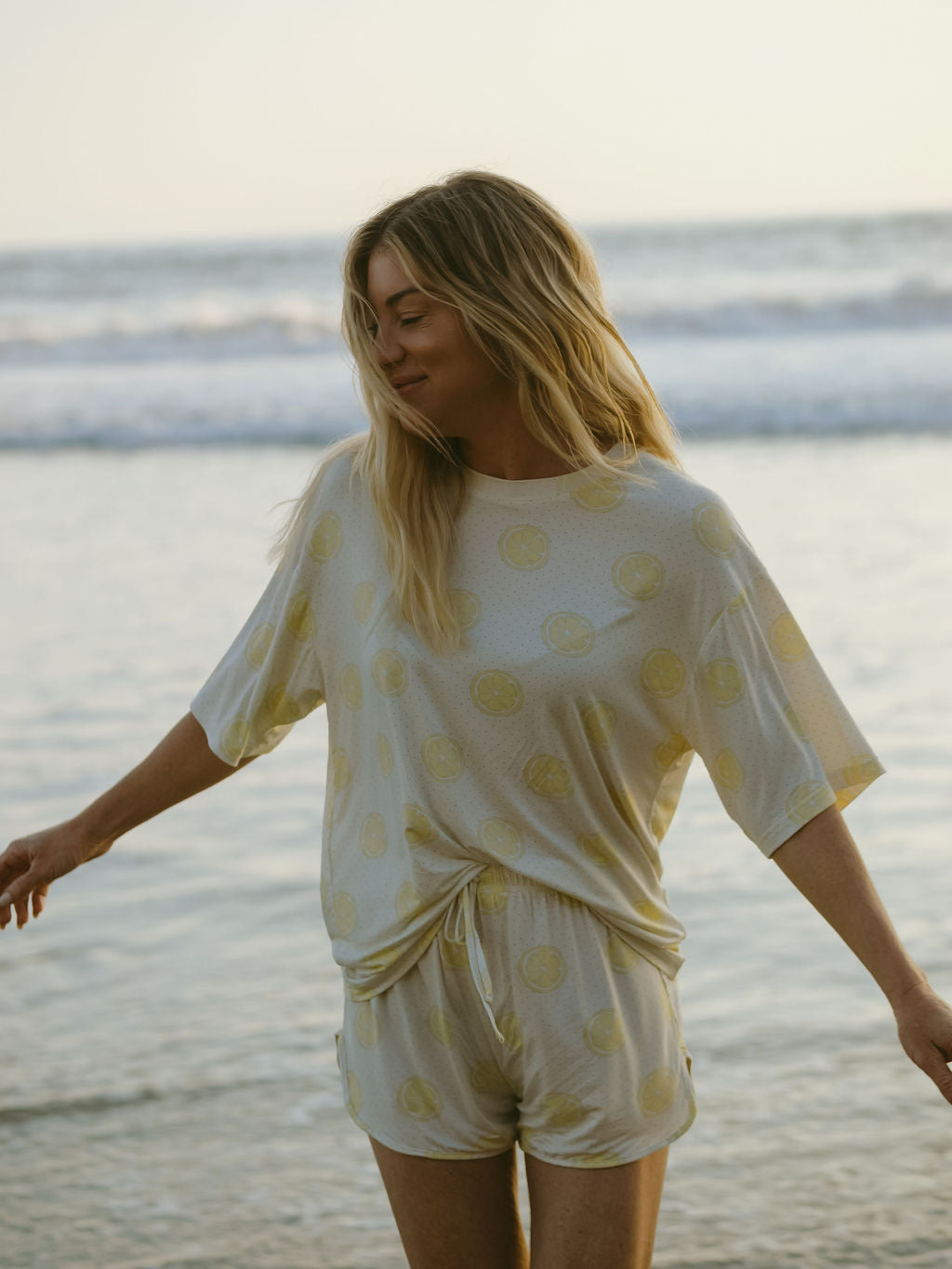 A woman with long blonde hair stands on a beach, smiling and wearing the forever french baby Women's Bamboo Short Set | Lemon Drop—a hypo-allergenic, breathable outfit with a cheerful yellow citrus fruit pattern.