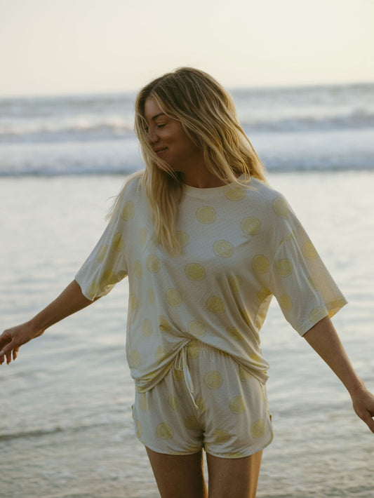A woman with long blonde hair stands on a beach, smiling and wearing the forever french baby Women's Bamboo Short Set | Lemon Drop—a hypo-allergenic, breathable outfit with a cheerful yellow citrus fruit pattern.