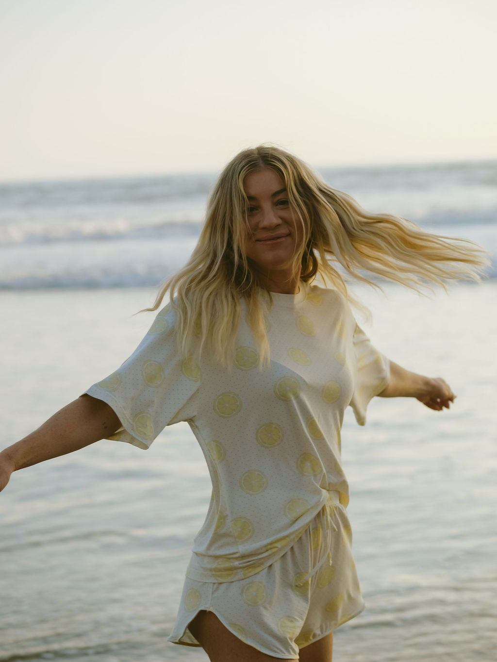 A woman with long blonde hair smiles and spins on the beach, wearing the Women’s Bamboo Short Set | Lemon Drop by forever french baby. The breathable set features a cheerful yellow pattern, with the ocean and waves in the background.
