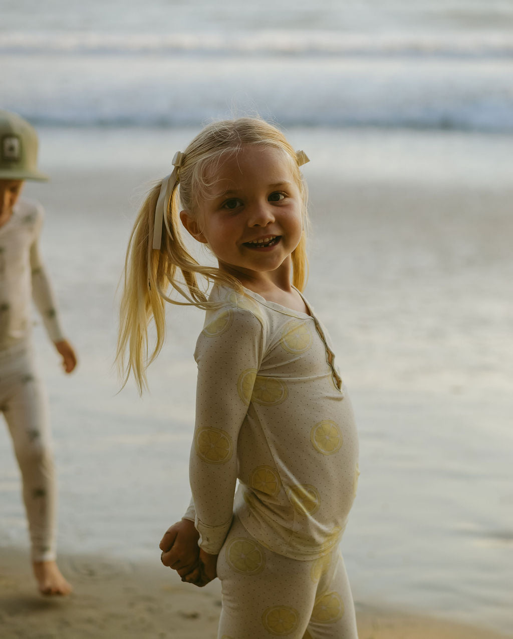 A young child with blonde pigtails smiles while standing on a sandy beach, wearing forever french baby's Bamboo Two Piece Pajamas in Lemon Drop. Another child appears partially in the background.