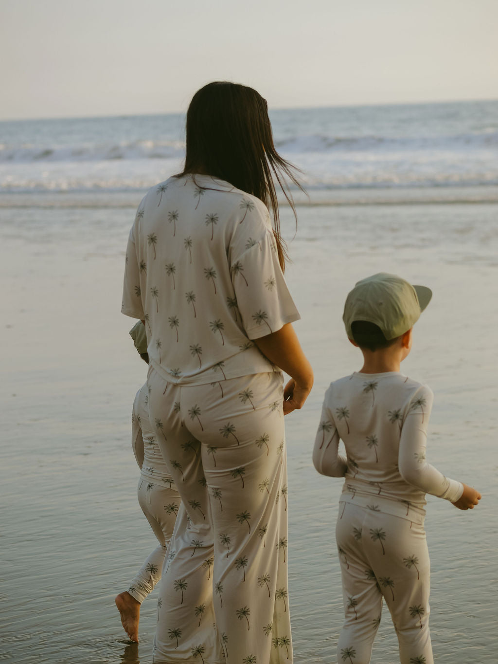 A woman and two children in matching forever french baby Women's Boxy Tee Lounge Sets with a palm tree pattern walk along the shoreline, the ocean and sky visible in the background.