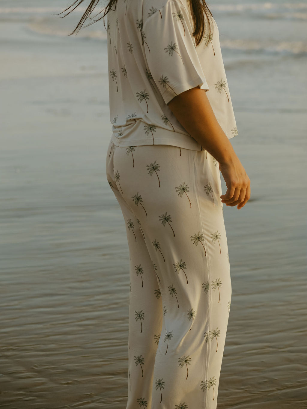 A woman stands on the beach in the forever french baby Women’s Boxy Tee Lounge Set | Palm Tree. The soft, hypo-allergenic bamboo set features a palm tree print, with ocean and sand behind her. Her face is not shown to highlight the outfit.