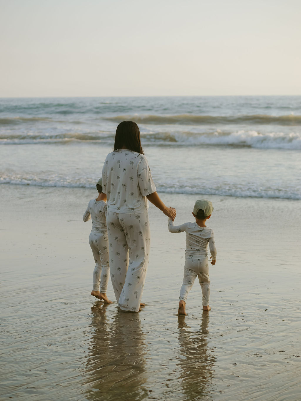 A woman and two children, all wearing forever french baby Women's Boxy Tee Lounge Set in Palm Tree, stroll along a sunlit beach toward the ocean with gentle waves rolling onto the shore.