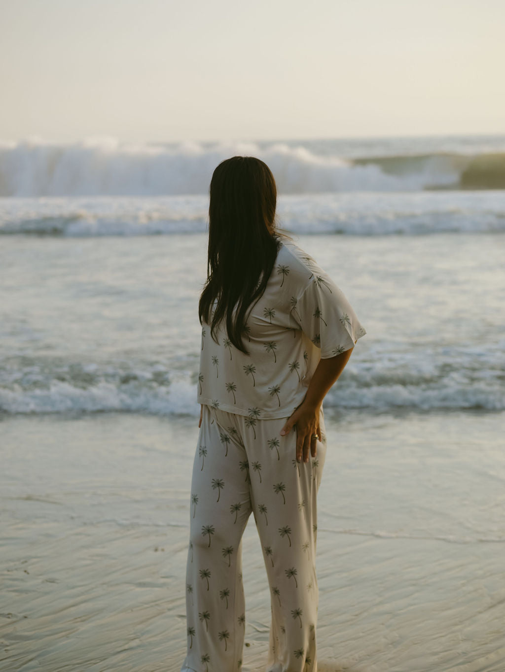 A woman with long dark hair, wearing the forever french baby Women's Boxy Tee Lounge Set in a palm tree print, stands on the beach facing the ocean waves at sunset.