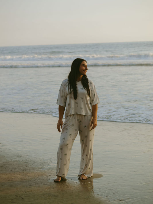 A woman wears the forever french baby Women's Boxy Tee Lounge Set in Palm Tree print, standing barefoot on a beach, smiling and gazing to the side as gentle waves roll up behind her.