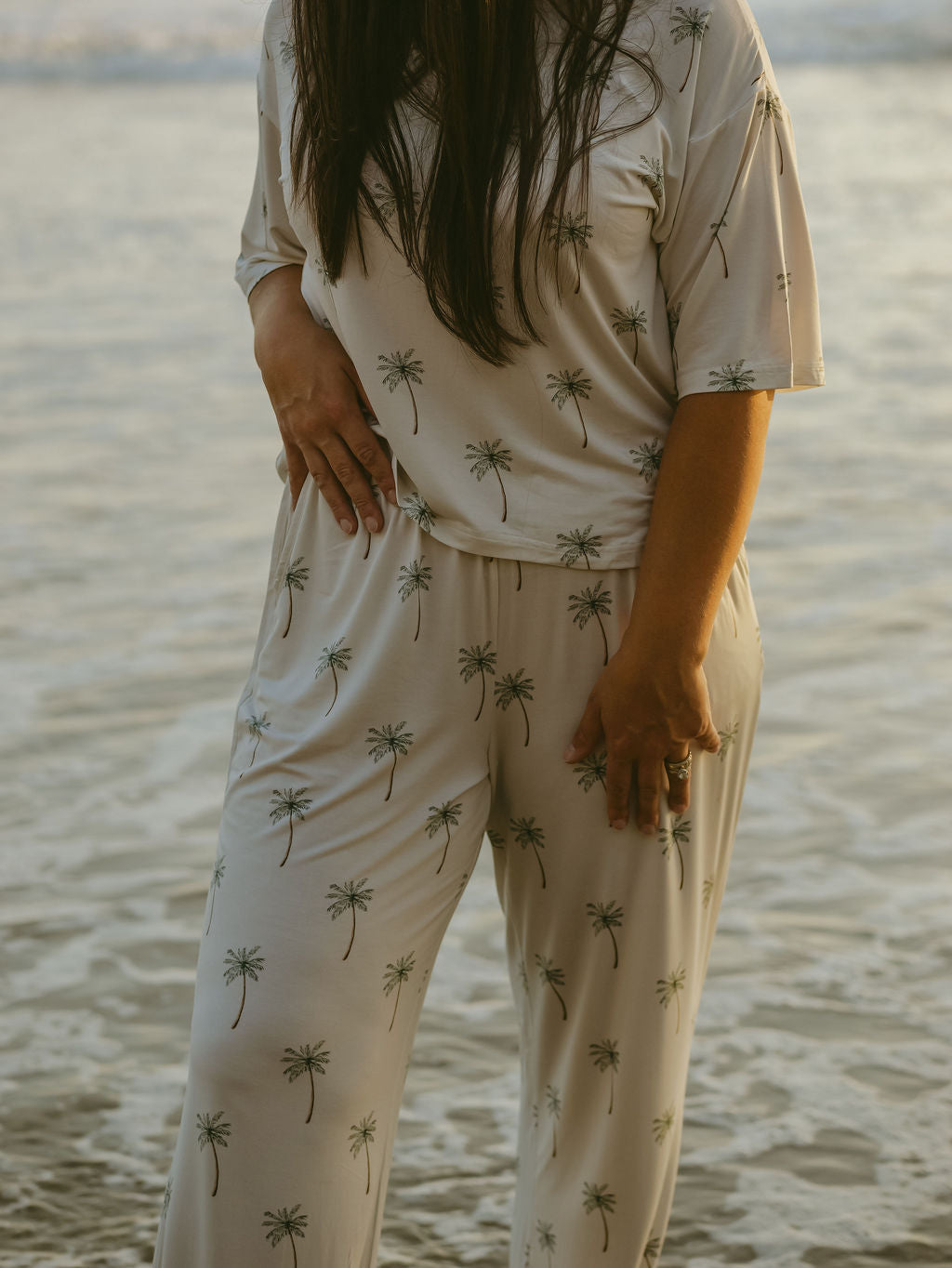 A person by the water wears the forever french baby Women's Boxy Tee Lounge Set | Palm Tree, a light-colored, hypo-allergenic outfit. The photo is cropped from the shoulders down.
