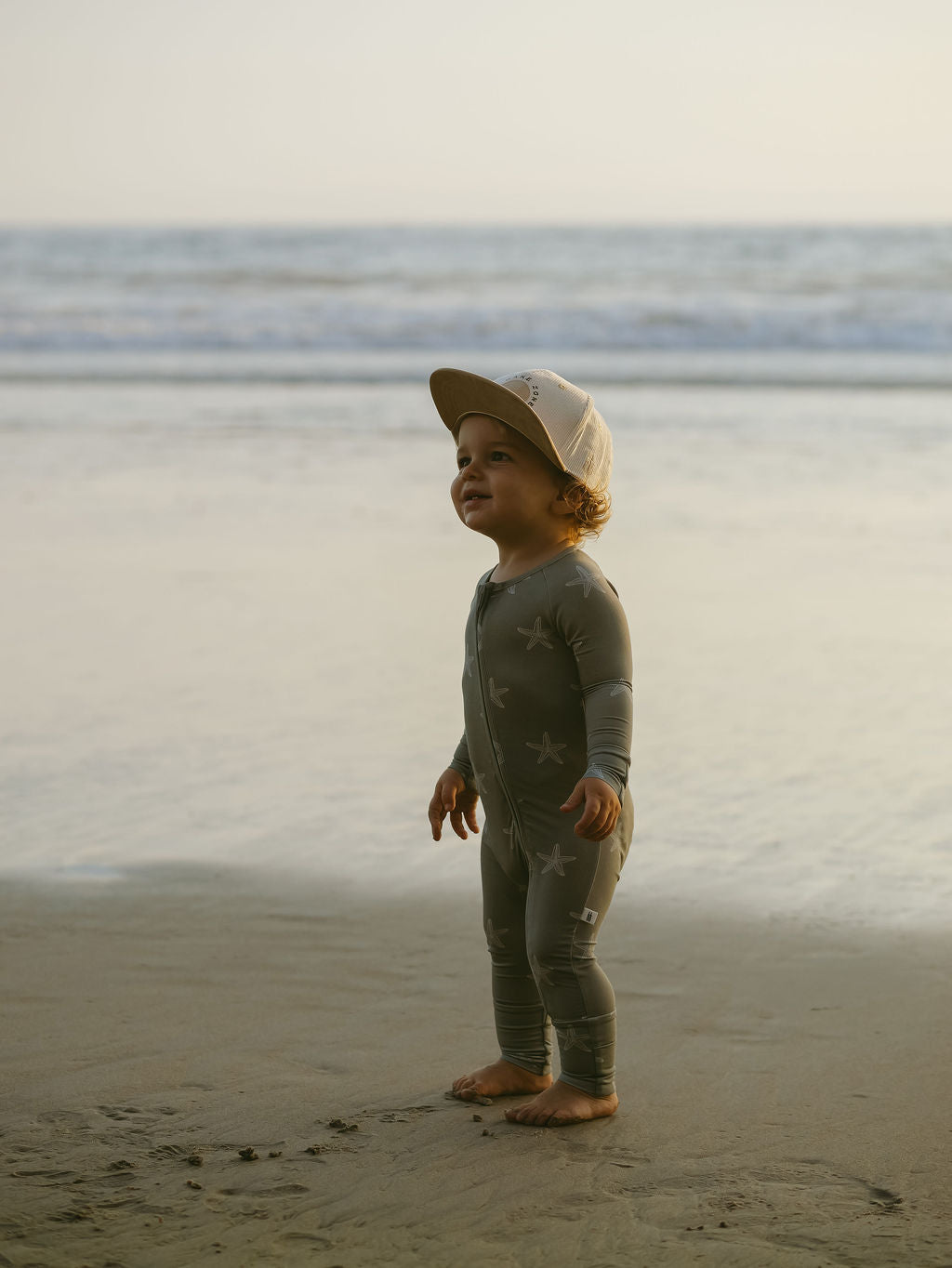A young child in forever french baby Bamboo Zip Pajamas | Starfish and a cap stands barefoot on a sandy beach, smiling up with the ocean and gentle waves behind.