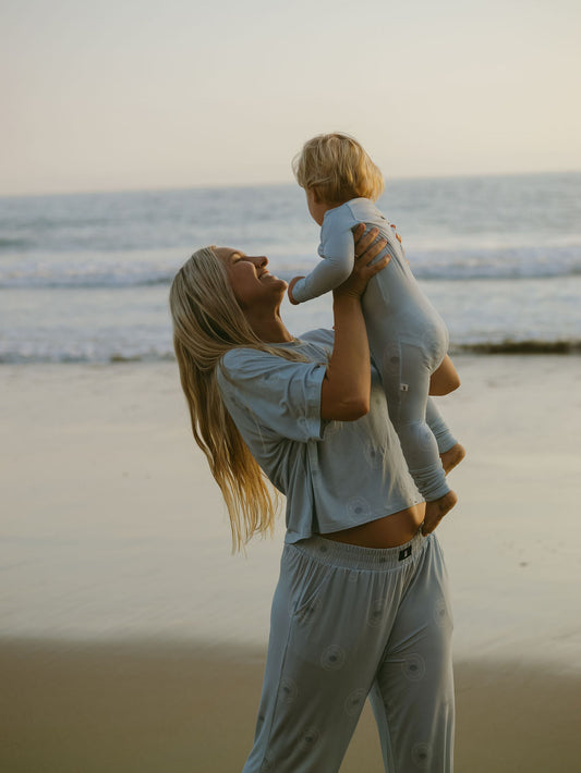 A woman in the Women's Boxy Tee Lounge Set | San Clemente by forever french baby lifts a toddler in matching pajamas on a sandy beach at sunset, with ocean waves in the background.