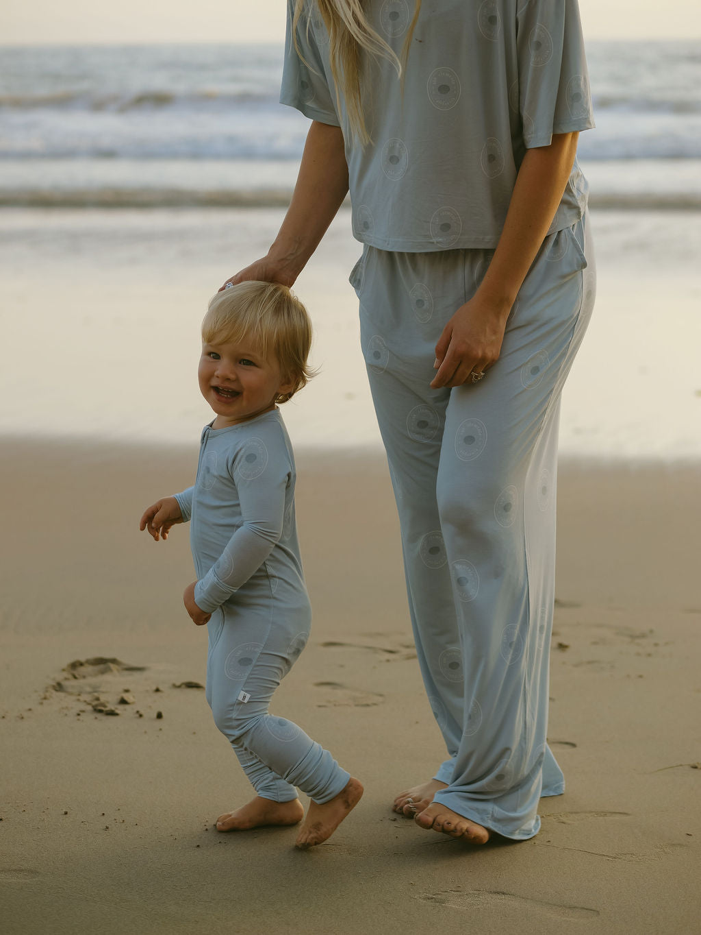 A small child in light blue Bamboo Zip Pajamas | San Clemente by forever french baby stands on the beach next to an adult, both wearing matching breathable sleepwear, with gentle ocean waves in the background.