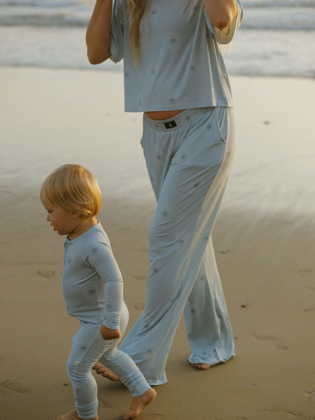 A woman and a toddler wear matching forever french baby Women's Boxy Tee Lounge Sets in San Clemente as they walk barefoot on a sandy beach near the water, gentle waves rolling in the background.