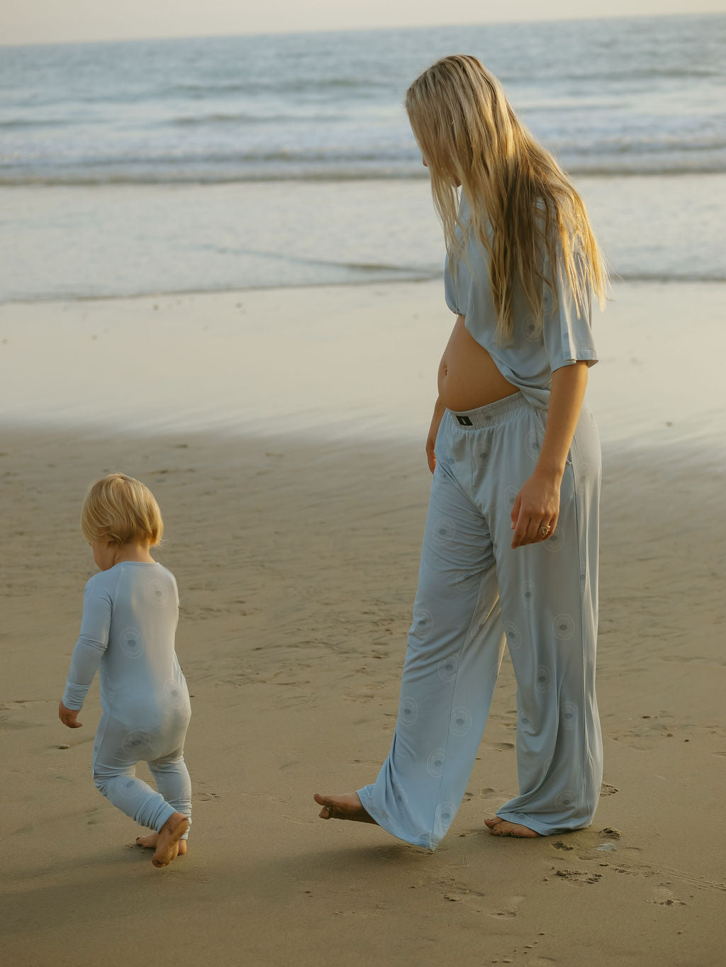 A pregnant woman and a young child, both wearing forever french baby’s Women's Boxy Tee Lounge Set in San Clemente, walk barefoot along the sandy beach at sunset as the woman looks down at the child beside her.