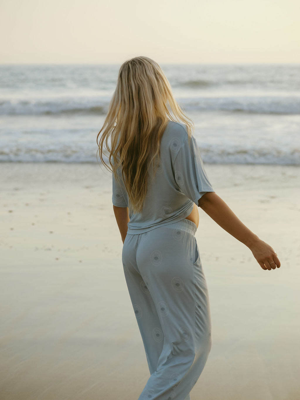 A woman with long blonde hair wears the forever french baby Women's Boxy Tee Lounge Set in San Clemente, standing on a beach facing the ocean under a soft golden sky.