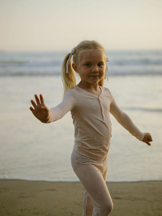 A young girl with blonde pigtails waves on a beach at sunset, wearing forever french baby Bamboo Two Piece Pajamas | Lobster—breathable children’s clothing that’s perfect for seaside evenings and gentle on sensitive skin.