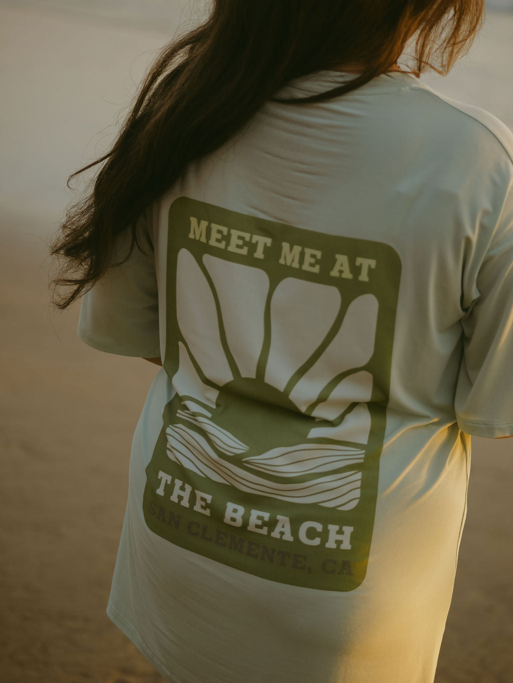 A woman with long brown hair stands on the beach wearing forever french baby's Short Sleeve Women's Bamboo Lounge Set, featuring a graphic on the back that says "Meet Me at the Beach, San Clemente, CA" with a sun and waves design.