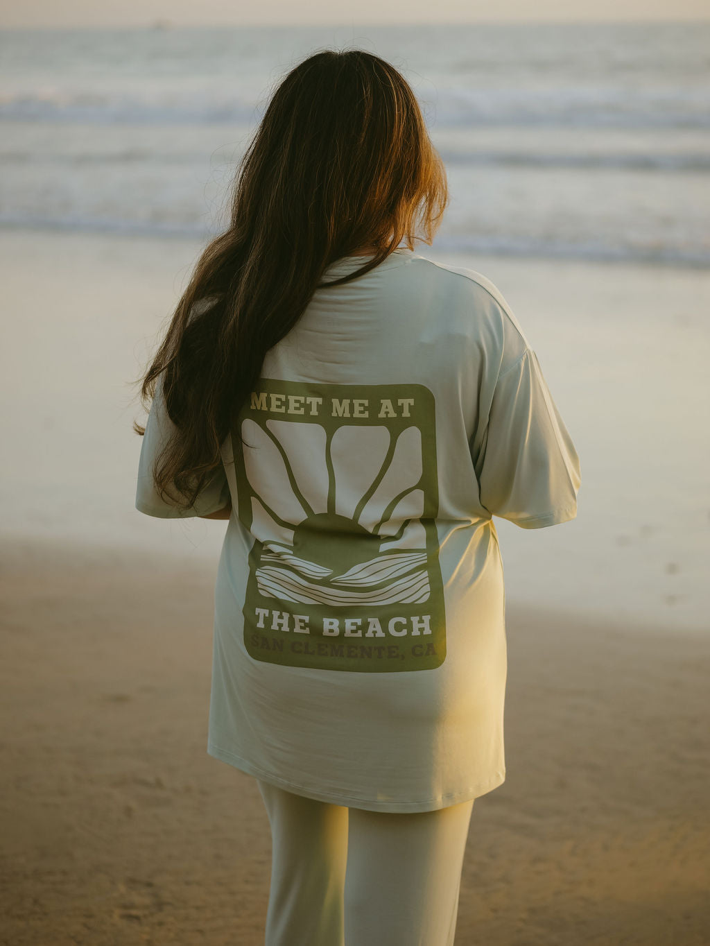 A person with long hair stands on the beach in the forever french baby Short Sleeve Women's Bamboo Lounge Set, featuring a sunrise graphic and “MEET ME AT THE BEACH, SAN CLEMENTE, CA” on the back.
