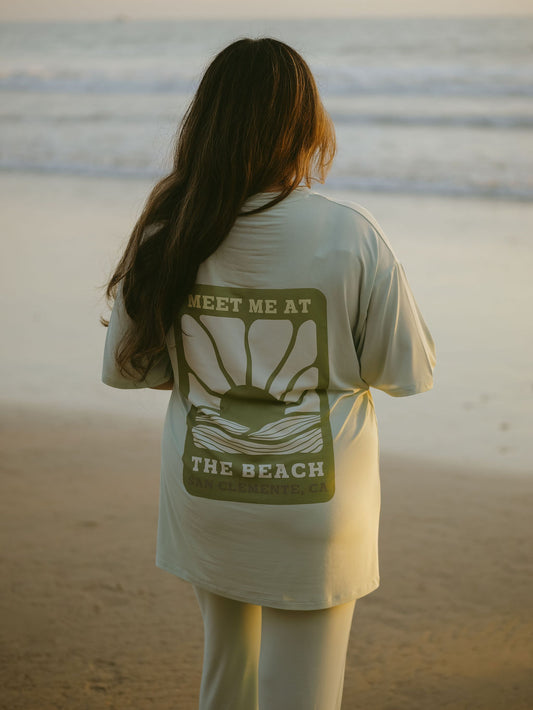 A person with long hair stands on the beach in the forever french baby Short Sleeve Women's Bamboo Lounge Set, featuring a sunrise graphic and “MEET ME AT THE BEACH, SAN CLEMENTE, CA” on the back.
