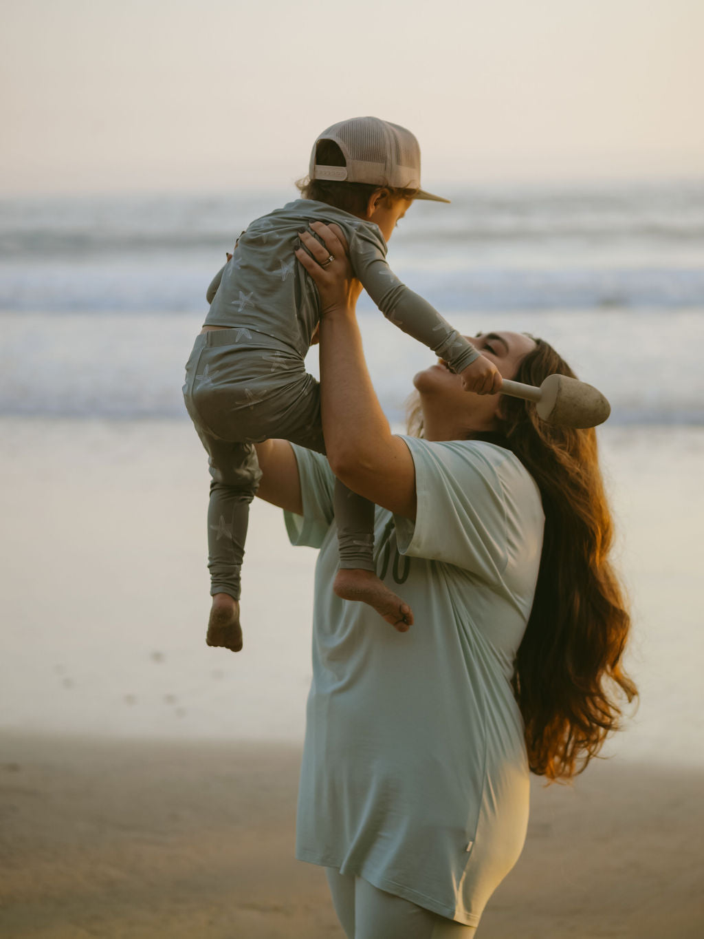 A joyful woman in the forever french baby Short Sleeve Women's Bamboo Lounge Set | Meet Me at the Beach lifts a child in matching bamboo pajamas at sunset by the ocean, with waves and soft light in the background.
