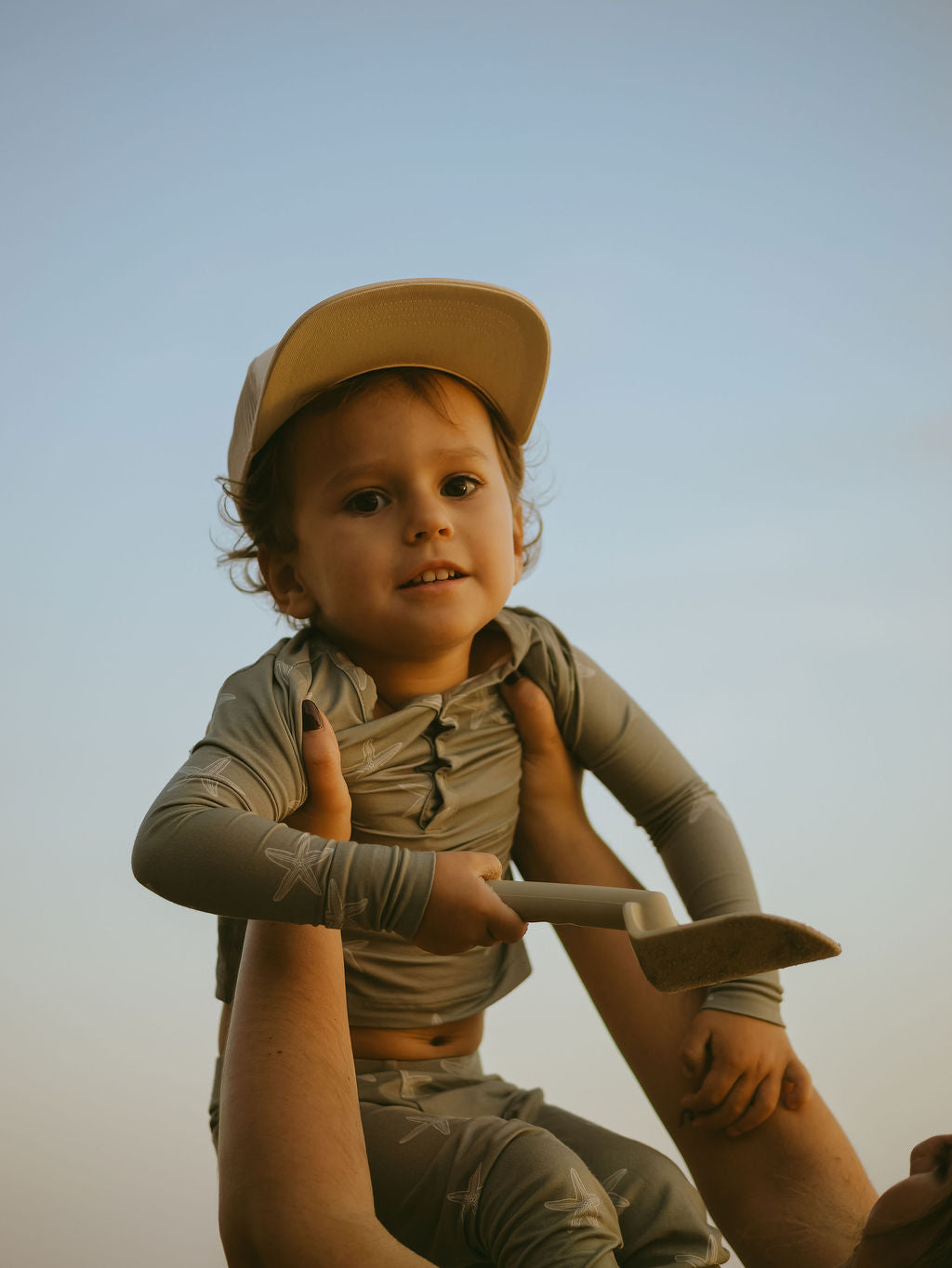 A young child in forever french baby's Bamboo Two Piece Pajamas | Starfish and a light-colored cap is held by an adult. The child smiles slightly, holding a small shovel against a clear sky background.