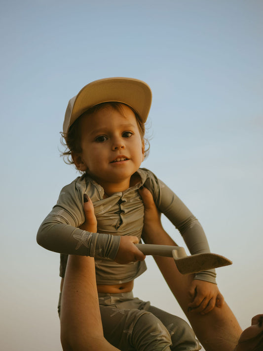 A young child in forever french baby's Bamboo Two Piece Pajamas | Starfish and a light-colored cap is held by an adult. The child smiles slightly, holding a small shovel against a clear sky background.