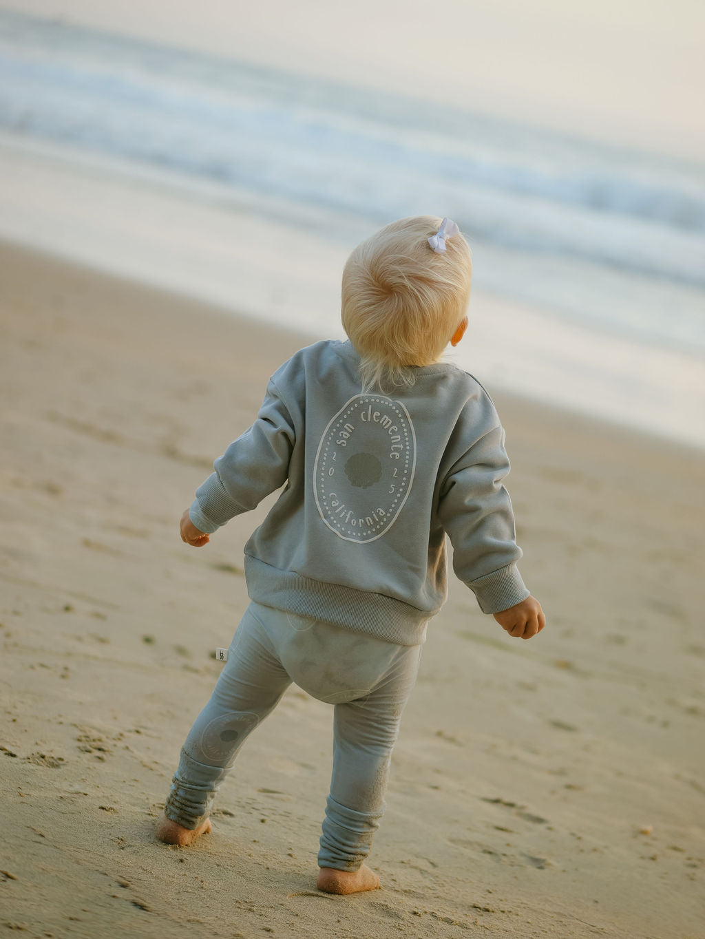 A young child in the forever french baby Child Sweatshirt | San Clemente stands barefoot on the sandy beach, gazing at ocean waves under a soft sky—a perfect coastal keepsake moment.