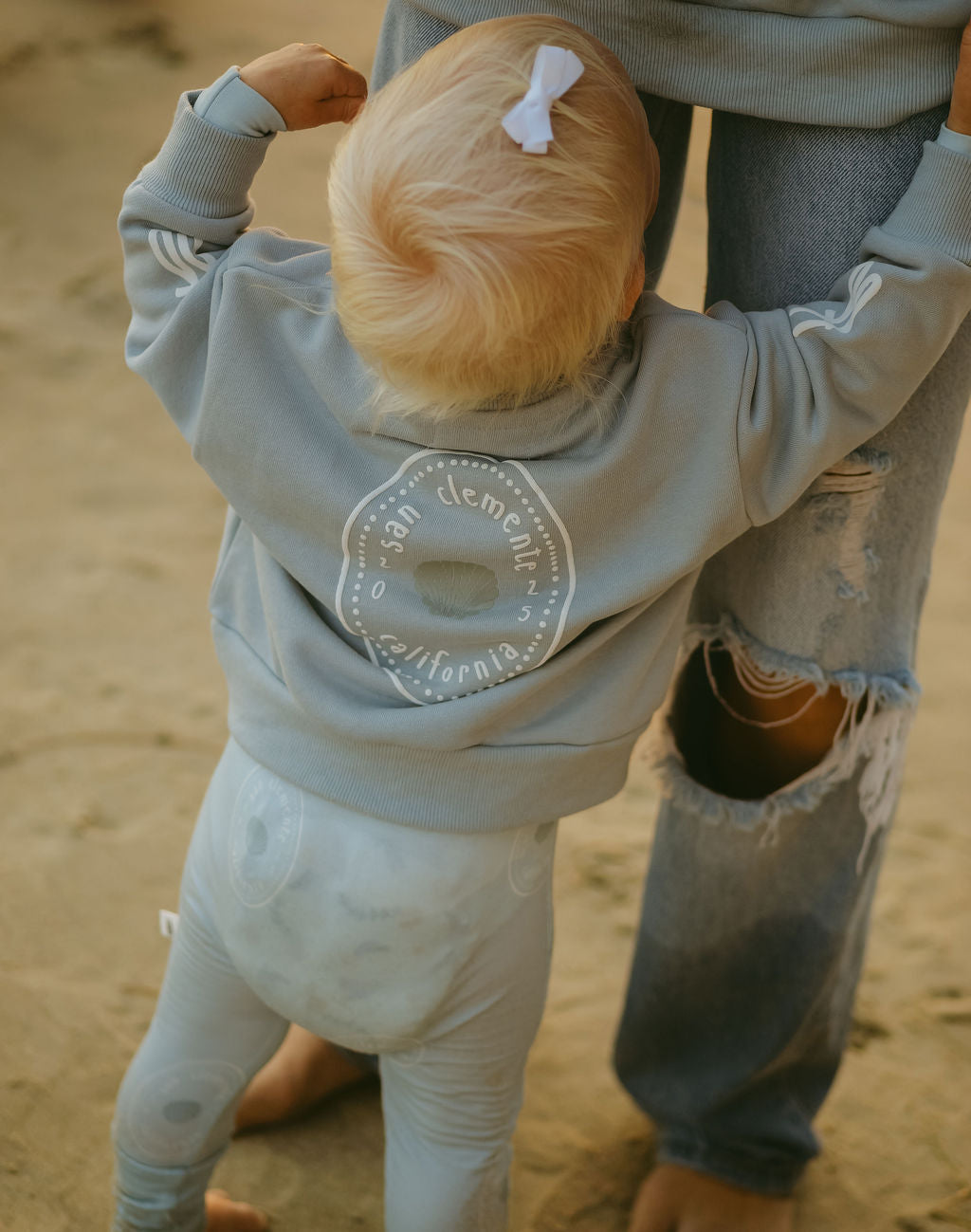 A blond toddler in a light blue Child Sweatshirt | San Clemente by forever french baby stands on the sand, clinging to an adult’s leg—a sweet moment captured in stylish California kids clothing.