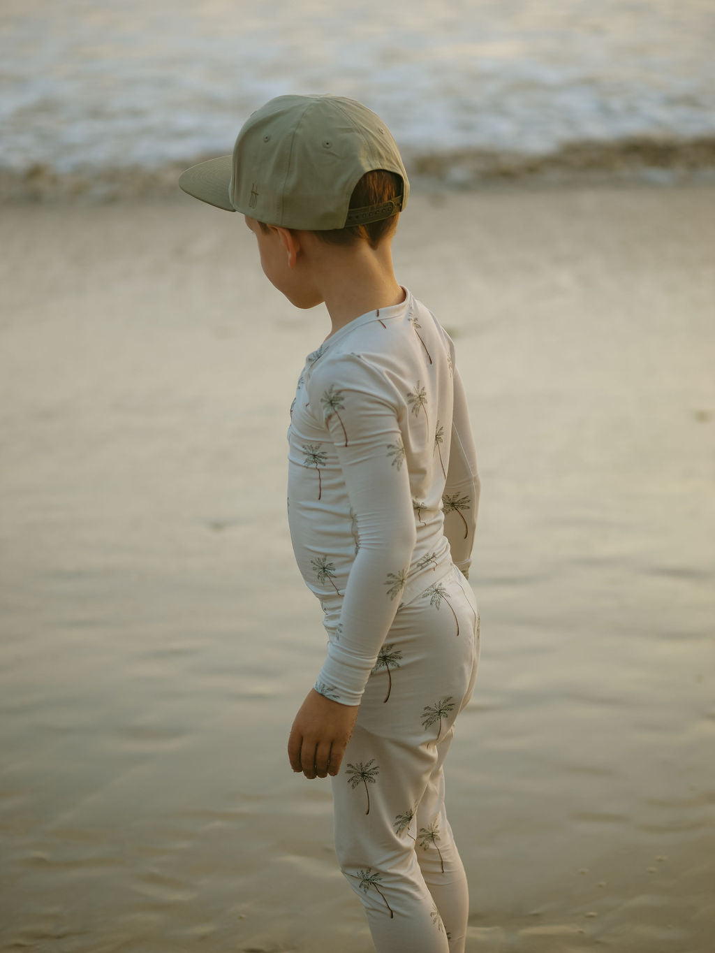 A young child wearing forever french baby's Bamboo Two Piece Pajamas in Palm Tree stands on wet beach sand, facing the ocean with a beige cap, captured from behind.