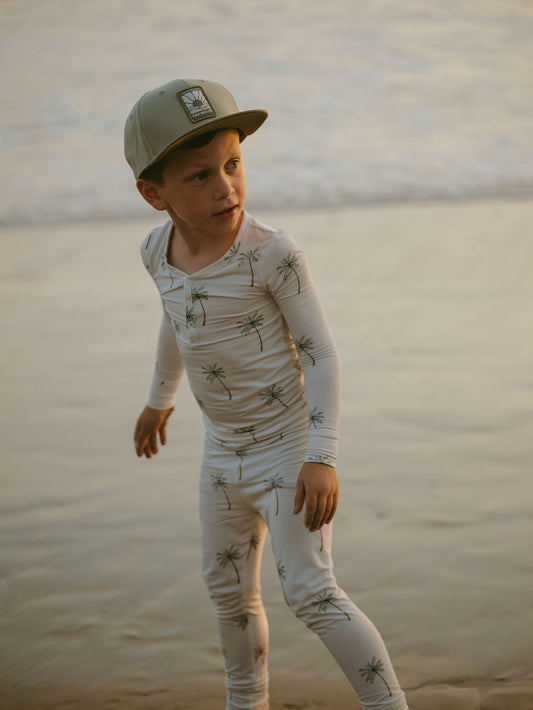 A young child wearing forever french baby’s Bamboo Two Piece Pajamas in a palm tree print, paired with a light green cap, stands barefoot on wet sand by the ocean at sunset.
