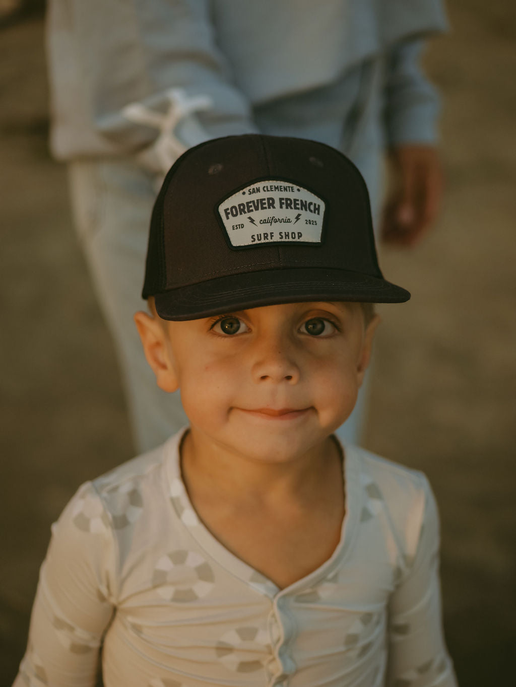 A young child wears forever french baby’s Bamboo Two Piece Pajamas | Lifesaver and a black "Forever French Surf Shop" hat, standing outdoors and looking at the camera while an adult hand rests on their shoulder.
