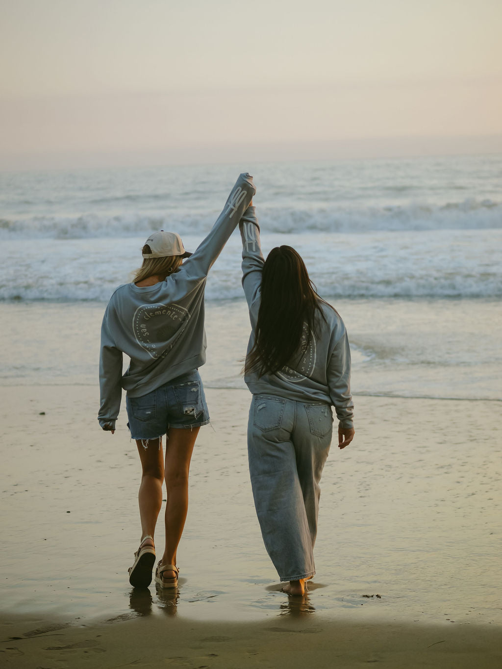 At sunset, two people stroll hand in hand along the beach, both wearing forever french baby’s Adult Sweatshirt | San Clemente—one pairs it with shorts and a cap, the other with loose pants—as waves softly roll in behind them.