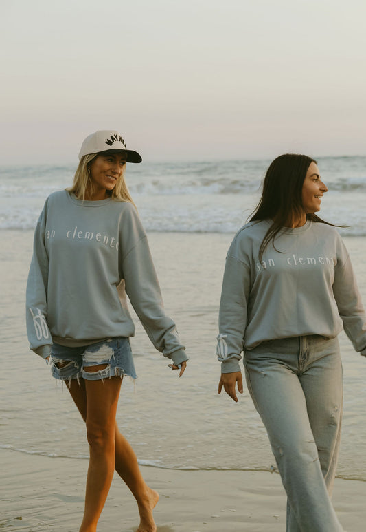 Two women wear forever french baby's Adult Sweatshirt | San Clemente as they stroll barefoot on the beach at sunset—one styles hers with denim shorts and a cap, the other with light jeans. Ocean waves shimmer behind them.