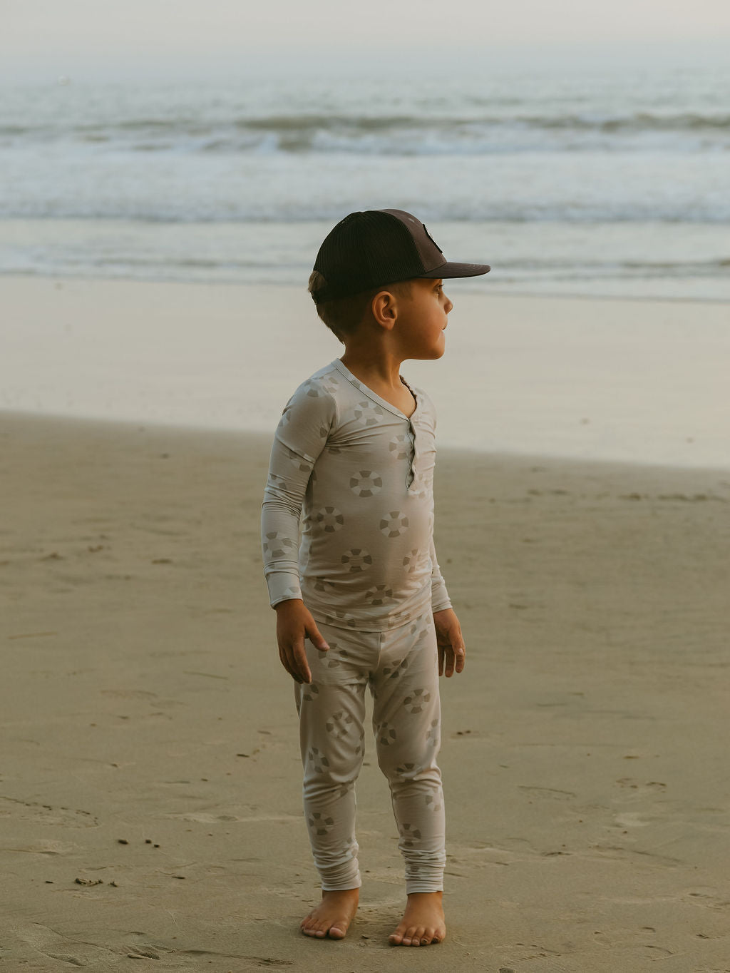 A young child wearing forever french baby’s Bamboo Two Piece Pajamas | Lifesaver and a black cap stands barefoot on the beach, gazing to the side as gentle ocean waves and a hazy sky fill the calm background.