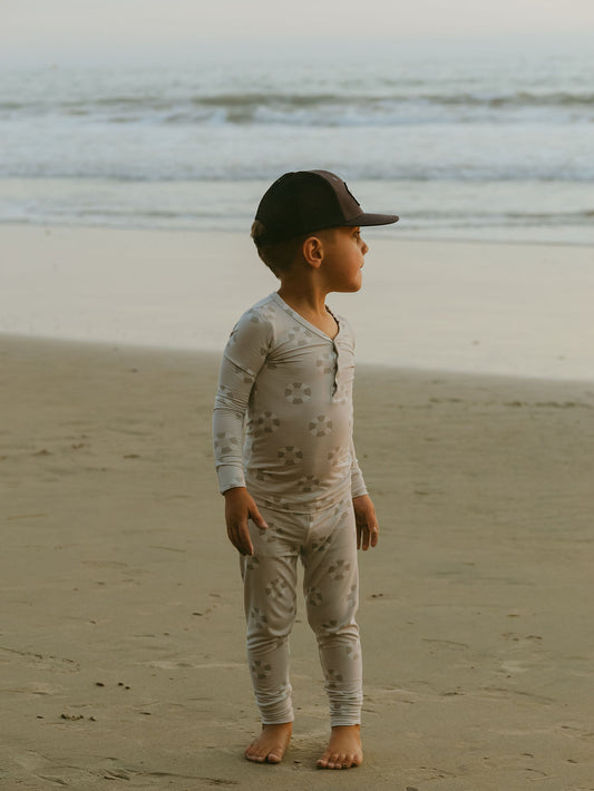 A young child wearing forever french baby’s Bamboo Two Piece Pajamas | Lifesaver and a black cap stands barefoot on the beach, gazing to the side as gentle ocean waves and a hazy sky fill the calm background.