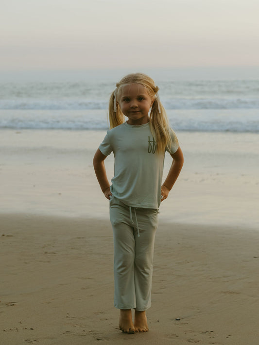 A young girl with blonde pigtails stands barefoot on the sand in a forever french baby Child Short Sleeve Bamboo Lounge Set | Meet Me at the Beach, with ocean waves and a pastel sky in the background.