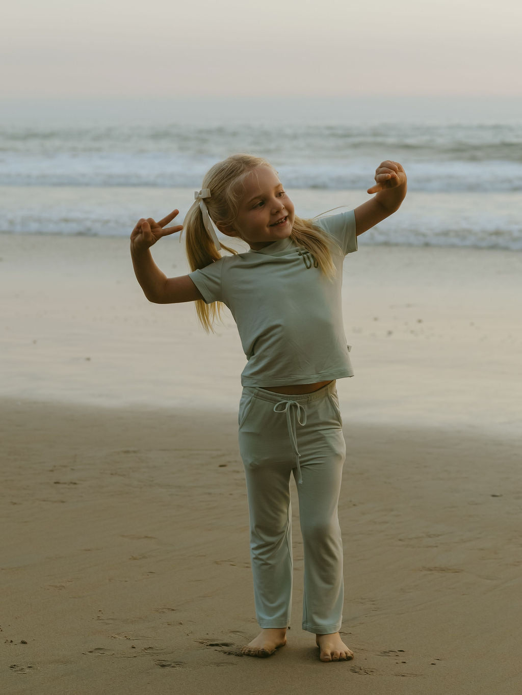 A young blonde girl smiles and makes peace signs barefoot on the beach, wearing the forever french baby Child Short Sleeve Bamboo Lounge Set | Meet Me at the Beach, with waves and a cloudy sky in the background.