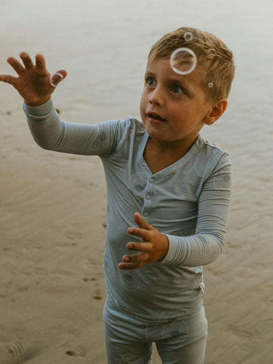 A young boy in forever french baby’s Bamboo Two Piece Pajamas | San Clemente, reaches up with excitement to catch floating bubbles on a sandy beach.